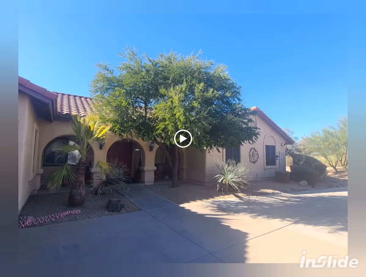 Exterior view of a single-story building with a tiled roof, arched entryways, and desert landscaping including palm trees and shrubs under a clear blue sky.