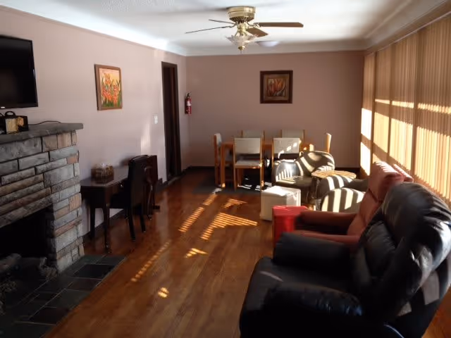 A cozy living room with wooden flooring, a stone fireplace on the left, a mounted flat-screen TV above the fireplace, and a small desk with a chair against the wall. There are two framed paintings on the walls. The room has a ceiling fan with a light fixture. At the far end, there is a dining table with four chairs. On the right side, there are large windows with vertical blinds letting in sunlight, and several cushioned chairs and a small ottoman arranged near the windows.
