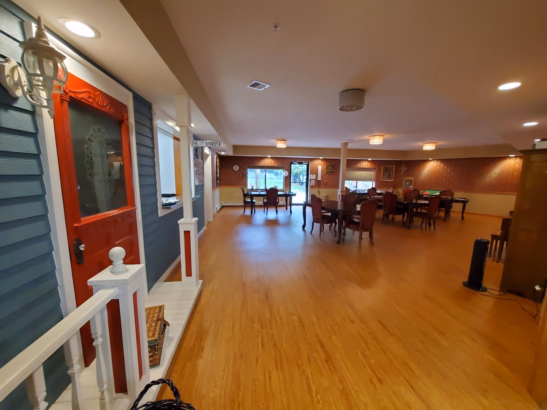 Interior view of a senior living facility common area with wooden flooring, several tables and chairs arranged for group activities or dining. On the left side, there is a decorative section resembling a small porch with a red door and blue siding. The room is well-lit with ceiling lights and has windows and a door at the far end providing natural light.