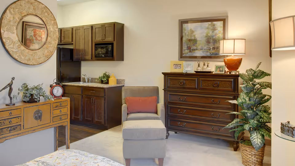 A cozy interior room featuring a small kitchenette with dark wood cabinets and a granite countertop, a comfortable gray armchair with a red cushion, a wooden dresser with framed photos and a lamp on top, a large leafy plant in a woven basket, and a decorative round mirror hanging above a wooden console table with plants and a clock.