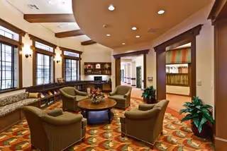 A spacious senior living facility lounge with curved chairs around a coffee table and plants.