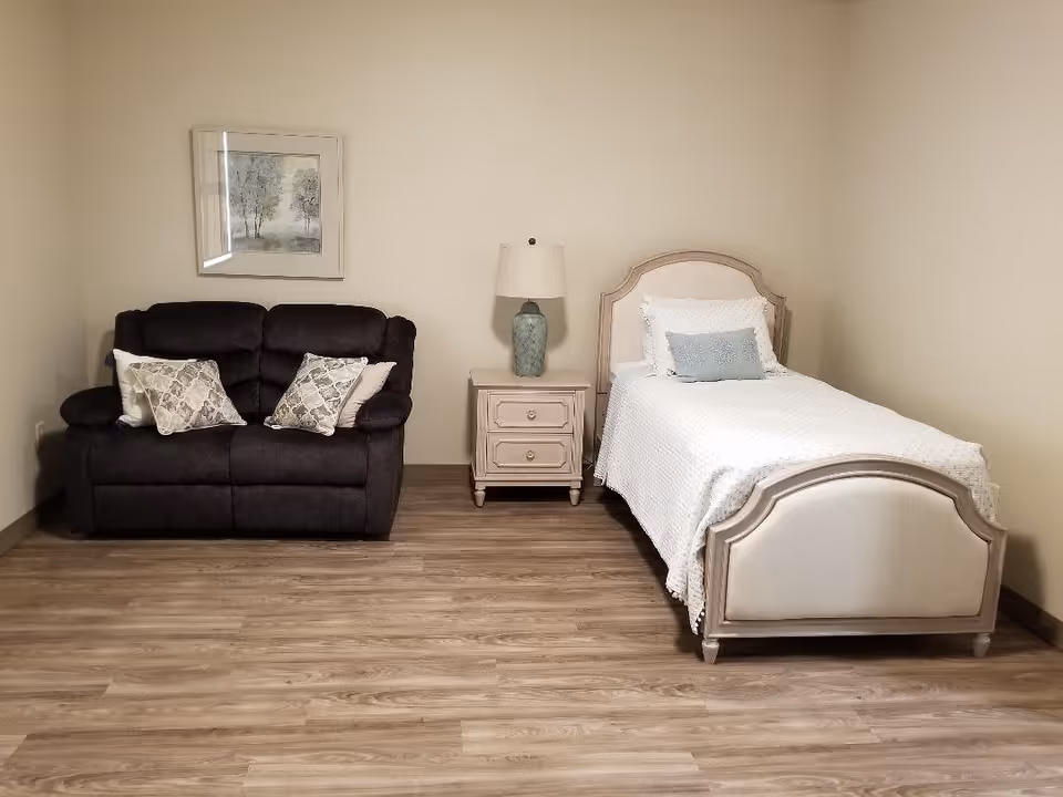 A simple bedroom with a single bed on the right side, covered with a white bedspread and two pillows. Next to the bed is a small nightstand with two drawers and a lamp with a blue base. On the left side of the room is a dark-colored loveseat with two patterned throw pillows. A framed picture of trees hangs on the wall above the loveseat. The floor is wooden, and the walls are painted a light beige color.