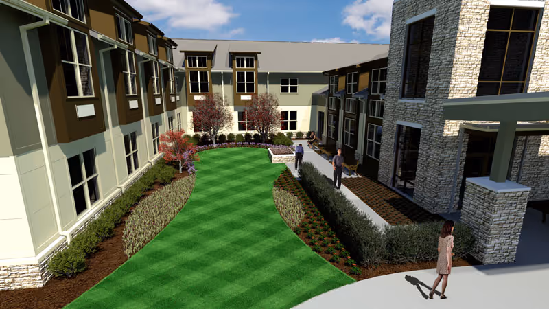 Outdoor courtyard area of a senior living facility with well-maintained green lawn, landscaped flower beds, and shrubs. The courtyard is surrounded by a multi-story building with large windows and stone accents. Several people are walking or standing on the paved pathways.
