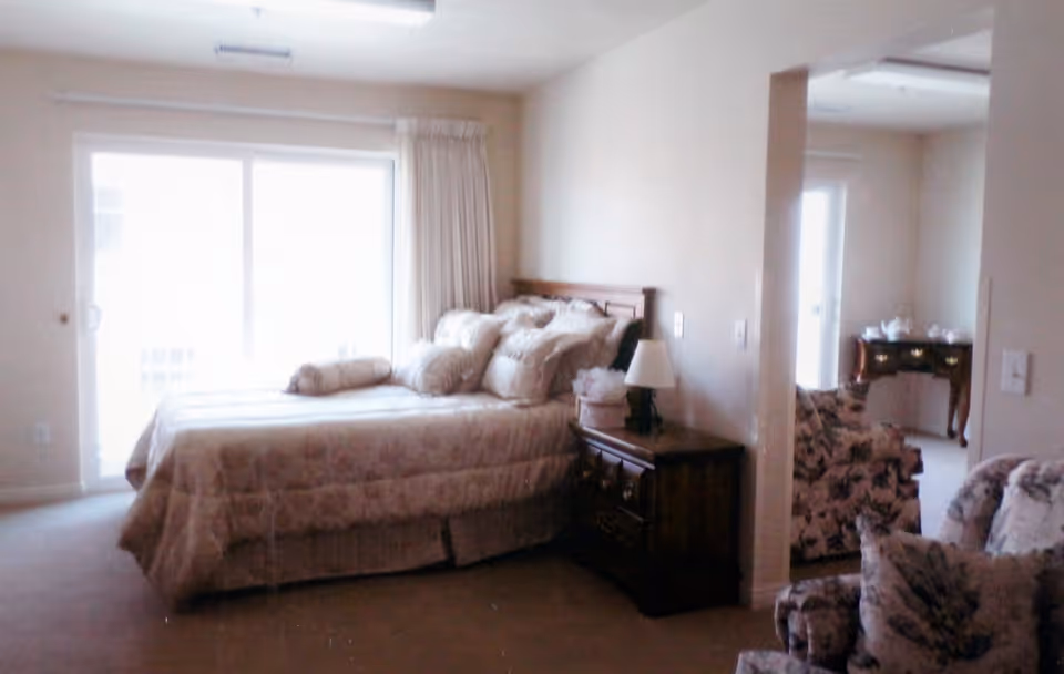 A bedroom with a large bed covered in a beige patterned comforter and multiple pillows. Next to the bed is a wooden nightstand with a lamp and a small decorative item. The room has beige walls and carpet, with a sliding glass door covered by curtains letting in natural light. Adjacent to the bedroom is a sitting area with floral upholstered chairs and a wooden table with tea set visible in the background.