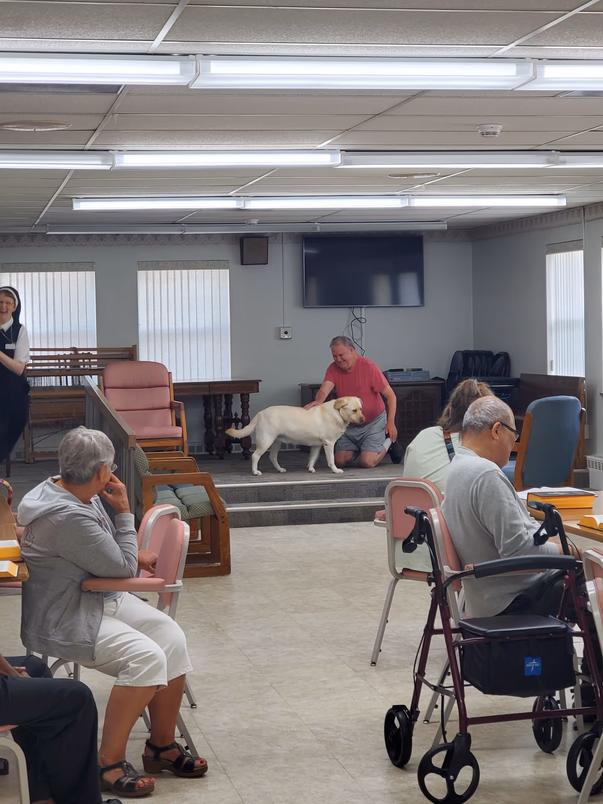 A group of elderly people seated in a common room with pink chairs and tables. A man in a red shirt kneels on a small stage petting a yellow Labrador retriever. A woman stands to the left smiling. The room has large windows with vertical blinds, a wall-mounted TV, and fluorescent ceiling lights.