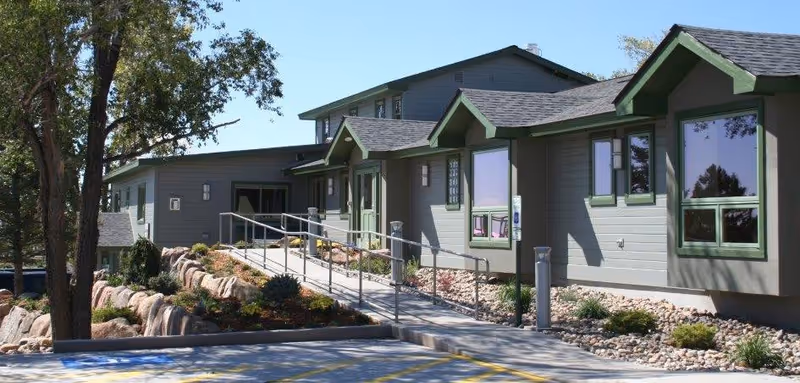 Exterior view of a single-story assisted living facility building with green trim around windows and doors, a wheelchair accessible ramp leading to the entrance, landscaped rocks and plants along the pathway, and trees in the background under a clear blue sky.