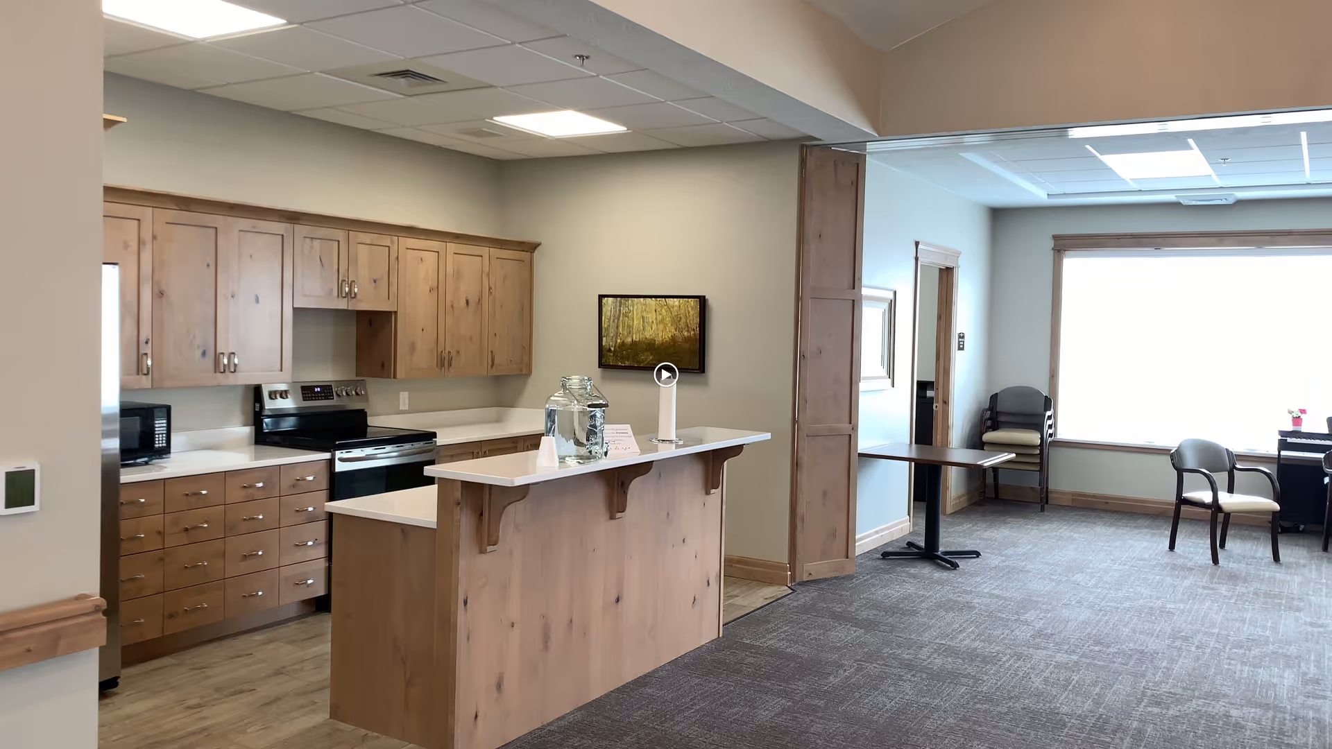 Interior view of a kitchen and adjacent common area in an assisted living facility. The kitchen features wooden cabinets, a stove, microwave, and a countertop island with a glass water dispenser. The adjacent area has carpeted flooring, a large window with natural light, a table, and several chairs.