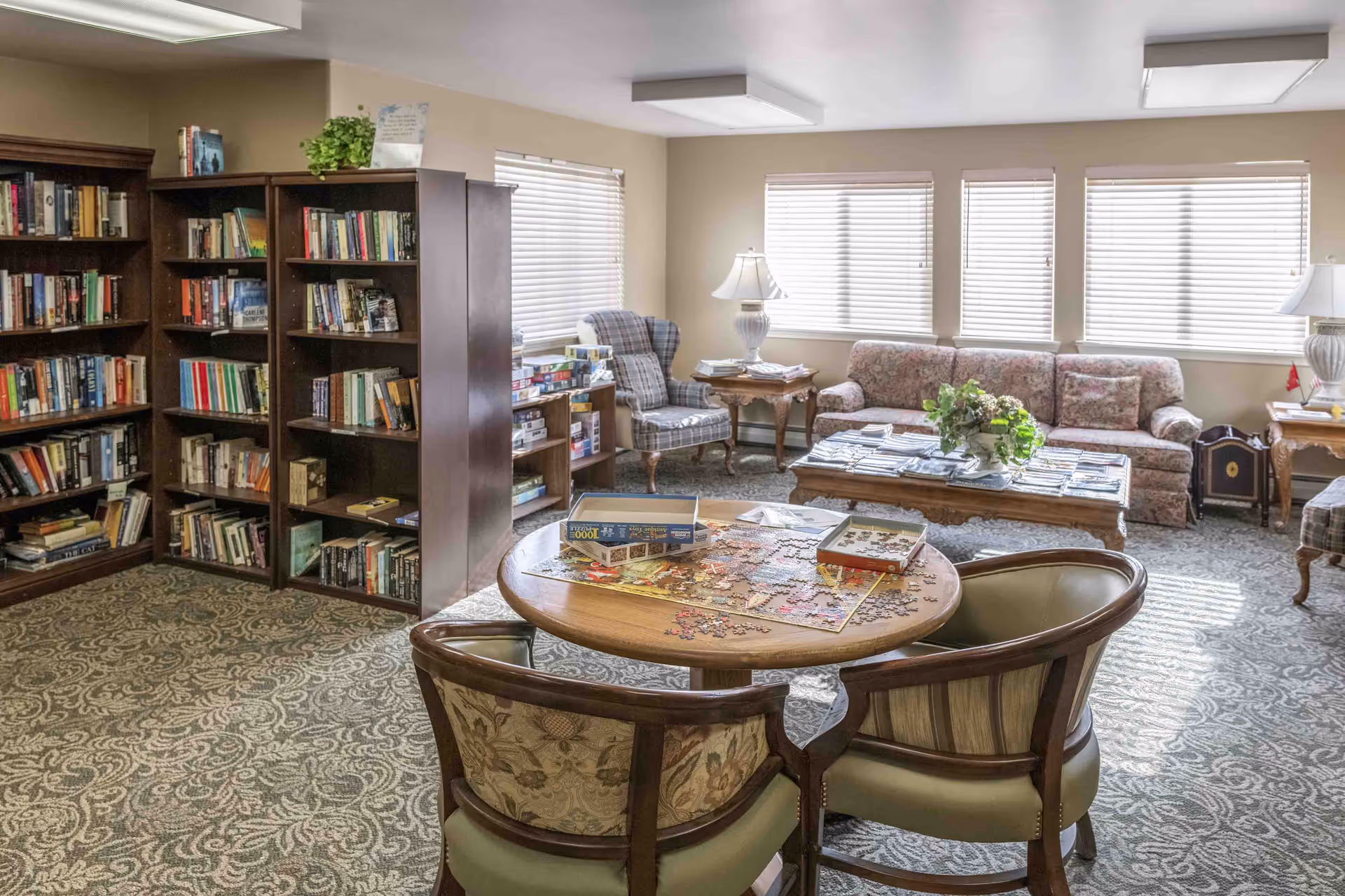 A cozy living room area with a round wooden table in the foreground featuring a partially completed jigsaw puzzle and puzzle box. Two upholstered chairs are placed around the table. In the background, there are bookshelves filled with books, a plaid armchair, a floral sofa, wooden side tables with lamps, and large windows with blinds letting in natural light. The room has patterned carpet flooring and a warm, inviting atmosphere.