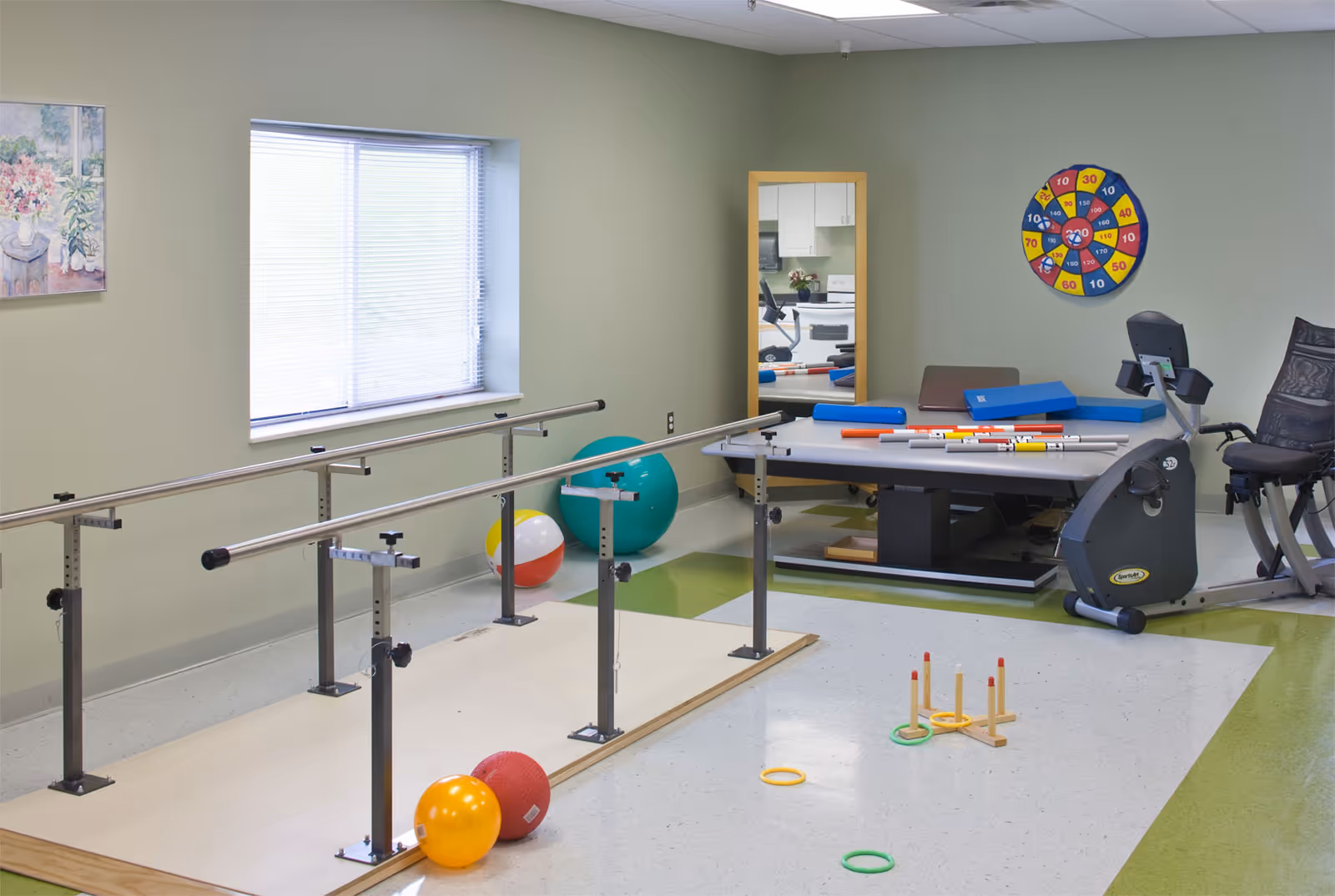 A therapy or exercise room with parallel bars for walking practice, various exercise balls, a stationary exercise bike, a table with foam rollers and other therapy equipment, a ring toss game on the floor, a large mirror on the wall, and a colorful dartboard target.