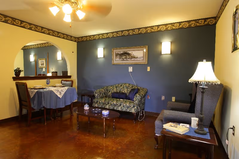 A cozy living room area with a floral patterned loveseat, a cushioned armchair, and a wooden coffee table. The walls are painted blue and beige with a decorative border near the ceiling. A framed picture hangs on the blue wall, flanked by two wall sconces. A ceiling fan with lights is above, and a small dining table with two chairs is set with a blue tablecloth and lace overlay near an arched opening in the wall. A table lamp with a fringed shade sits on a side table next to the armchair.