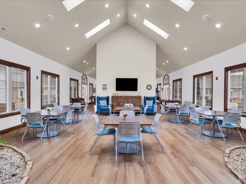 Bright and spacious common area with high vaulted ceilings and skylights. The room features multiple tables with chairs arranged for dining or socializing, two blue armchairs with patterned cushions, a large flat-screen TV mounted on the wall above a decorative wooden cabinet, and large windows with blinds letting in natural light. The floor is wood with small indoor garden areas filled with rocks on either side.