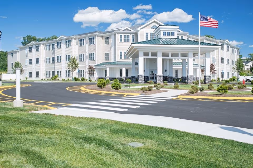 Exterior view of a large, three-story assisted living facility with white siding and green roofs under a blue sky with scattered clouds. The building has multiple windows and a covered entrance with stone pillars. An American flag is flying on a flagpole near the entrance. There is a paved driveway with a crosswalk and landscaped greenery in the foreground.