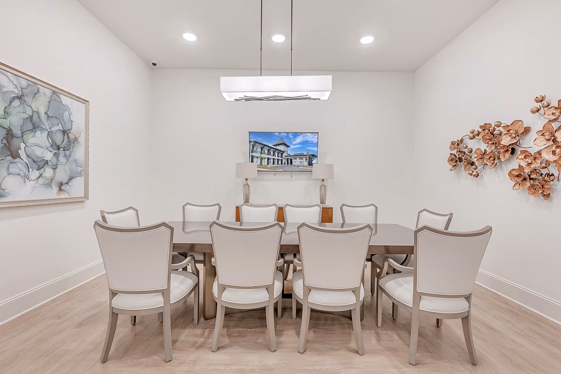 A modern dining room with a rectangular wooden table surrounded by ten white cushioned chairs. The room features light wood flooring, white walls, a large abstract painting on the left wall, and a decorative metal floral wall art on the right. A flat-screen TV mounted on the far wall displays an image of a building under a blue sky. Two table lamps with beige shades sit on a wooden console beneath the TV. A rectangular ceiling light fixture hangs above the table.
