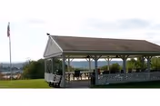 A covered outdoor pavilion with a pitched roof and open sides, containing several tables and chairs. The pavilion is situated on a grassy area with a flagpole nearby and a scenic view in the background.
