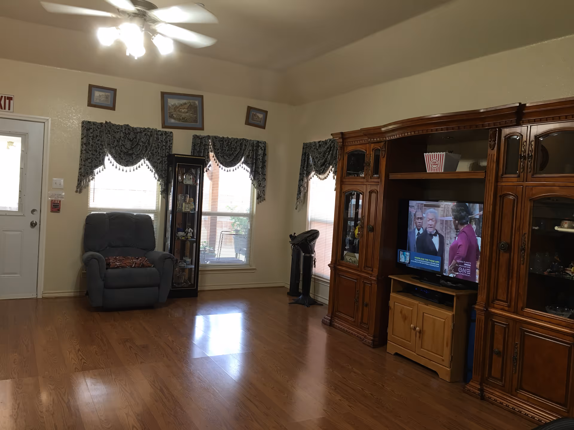 A living room with wooden flooring, a ceiling fan with lights, a gray recliner chair near two windows with patterned valances, a glass display cabinet, and a large wooden entertainment center with a television showing a scene from a TV show or movie.