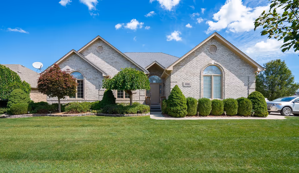 Front exterior view of a single-story brick building with a well-maintained lawn and various shrubs and trees under a partly cloudy blue sky.