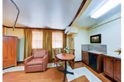 A cozy interior room featuring a single armchair, a round wooden table with two chairs, a wooden cabinet with a built-in fireplace, and a framed picture on the wall. The room has two windows with beige curtains and wooden flooring.