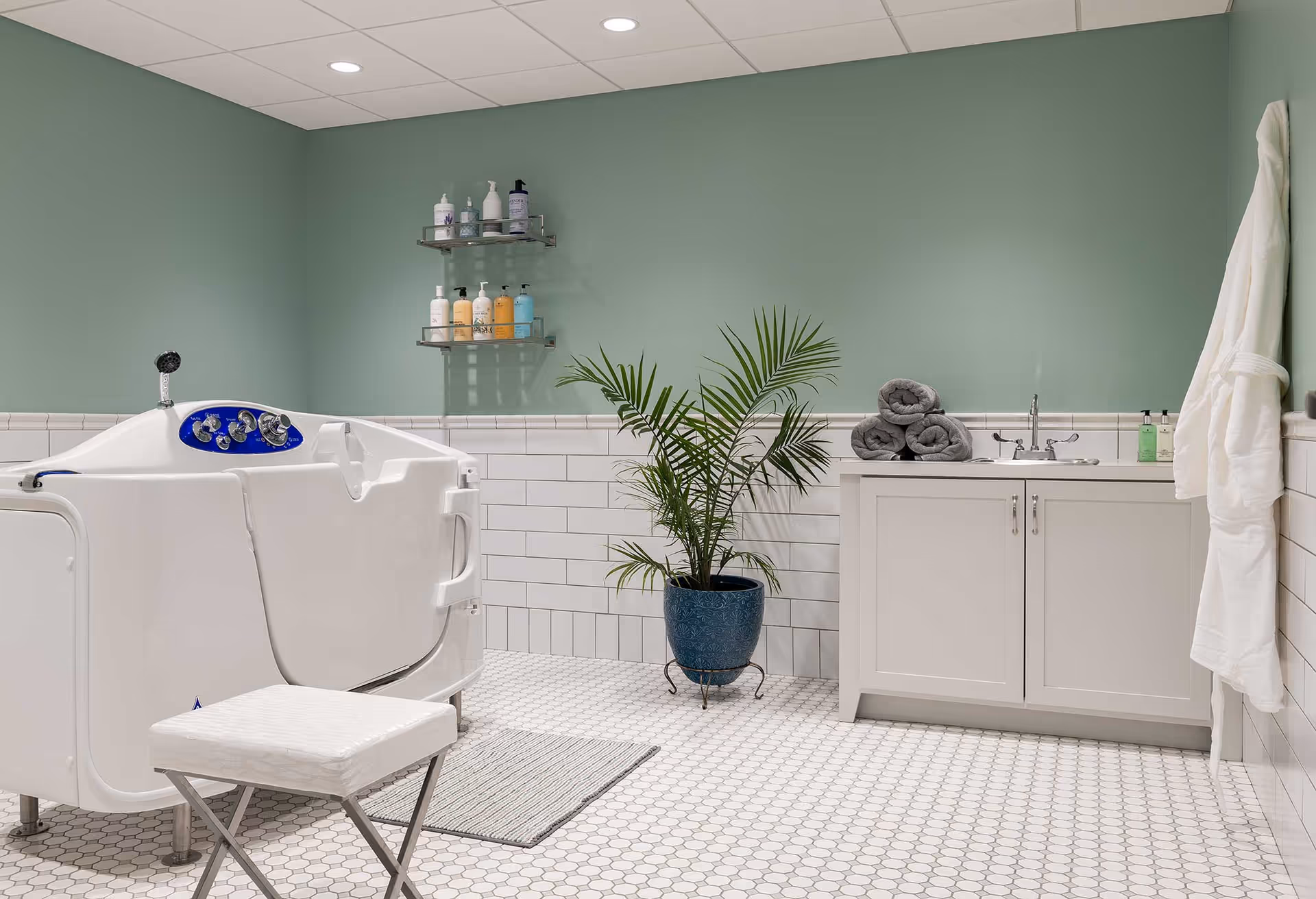 A clean bathing room featuring a walk-in tub, sink cabinet with folded towels, a potted plant, and a stool against a green wall.
