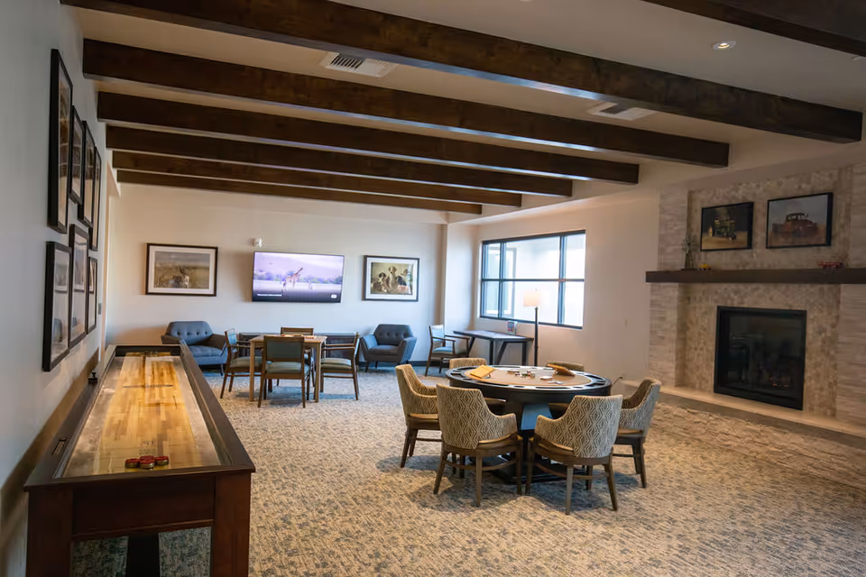 Spacious senior living lounge with a shuffleboard table, round dining table, chairs, and a fireplace.