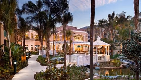 Courtyard with palm trees, a white gazebo by a pond and winding walkways leading to a multi-story building at dusk.