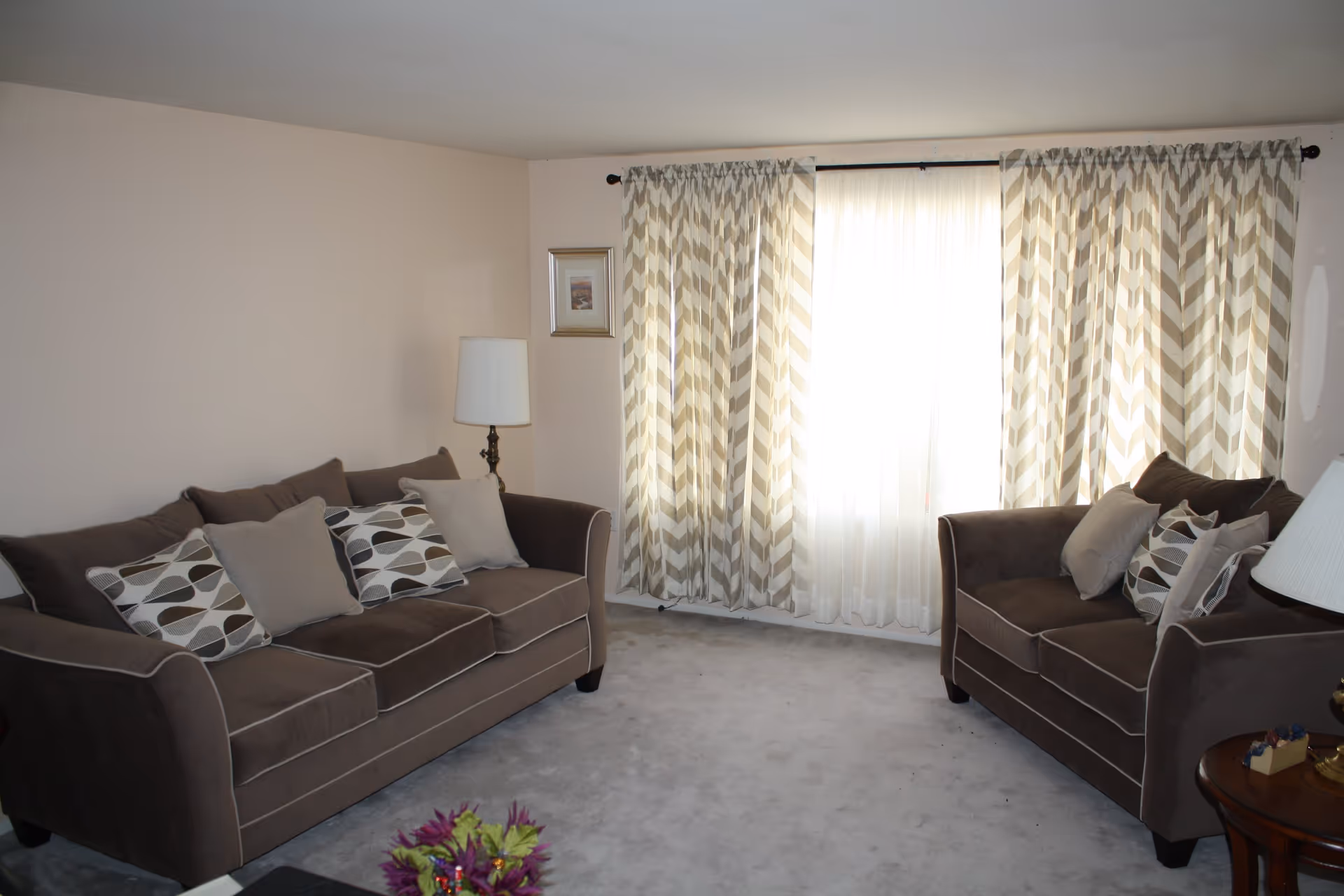Bright living room with two brown sofas facing a window with patterned curtains, throw pillows, a floor lamp and a small side table.