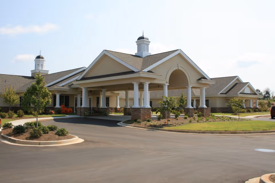 Exterior view of a single-story senior living facility building with a covered entrance supported by white columns and a landscaped driveway with small trees and shrubs.