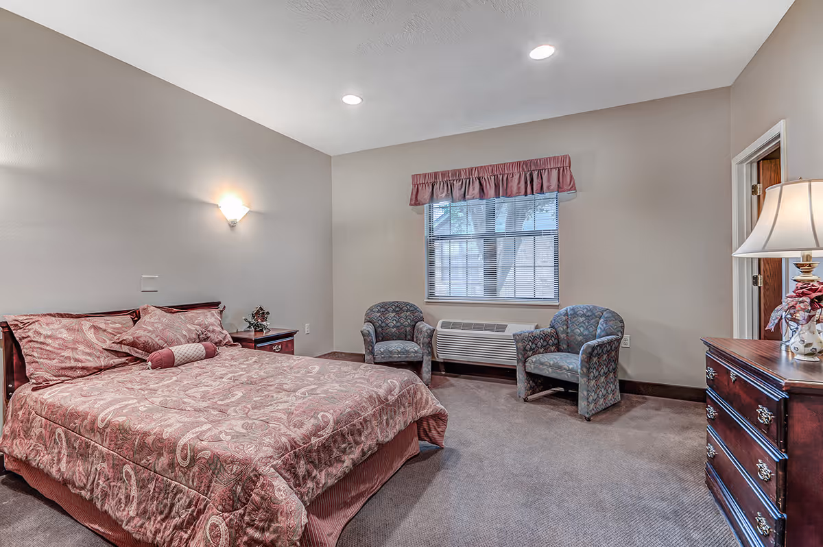 Furnished bedroom with a patterned double bed, two armchairs by a window, and wooden dressers.
