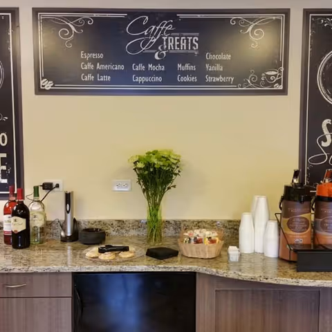 A coffee and treats station with a granite countertop featuring bottles of wine, a vase with green flowers, a basket of assorted snacks, disposable cups, and coffee dispensers. Above the counter is a blackboard-style menu listing espresso, caffe americano, caffe latte, caffe mocha, cappuccino, muffins, cookies, chocolate, vanilla, and strawberry.