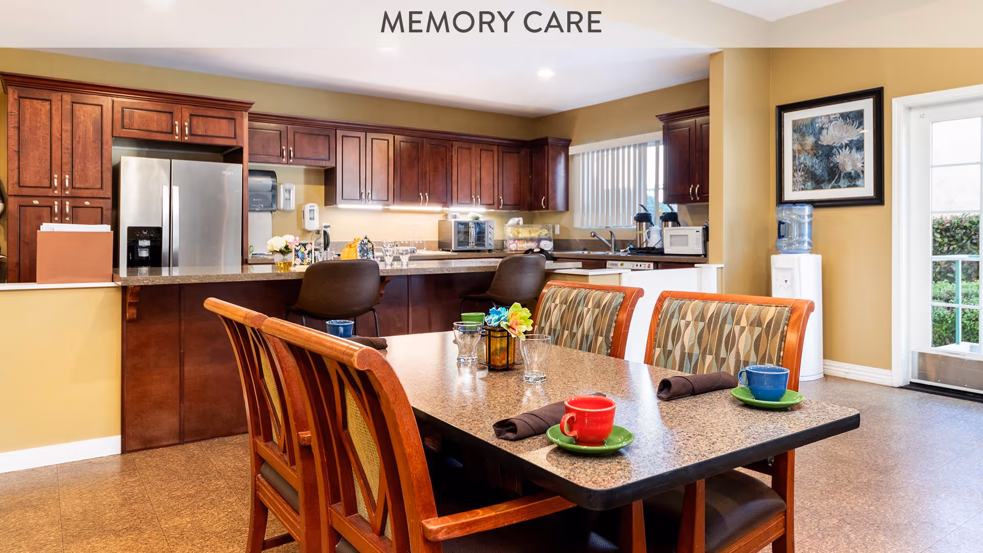 A bright and clean kitchen and dining area in a memory care facility. The kitchen features dark wood cabinets, a stainless steel refrigerator, and various appliances. In the foreground, there is a dining table with four chairs, each set with colorful cups and napkins. A water dispenser and framed artwork are visible near a door leading outside.