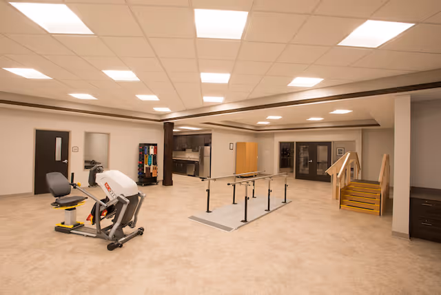 Spacious rehabilitation room with exercise equipment, parallel bars, and a practice staircase inside a senior care facility.