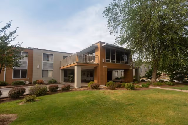 Exterior view of a two-story building with a covered entrance and balcony, surrounded by green grass, bushes, and trees under a cloudy sky.