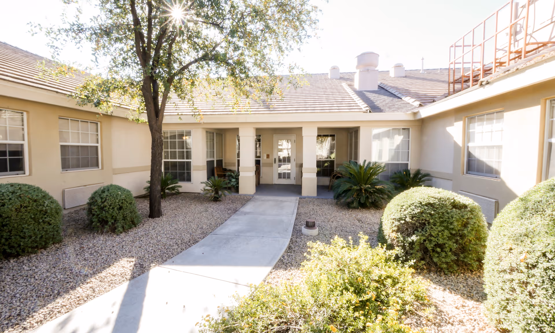 Outdoor courtyard area of a senior living facility with a concrete walkway leading to a covered entrance. The courtyard is landscaped with bushes, small trees, and gravel ground cover. The building has beige walls and multiple windows with white frames.