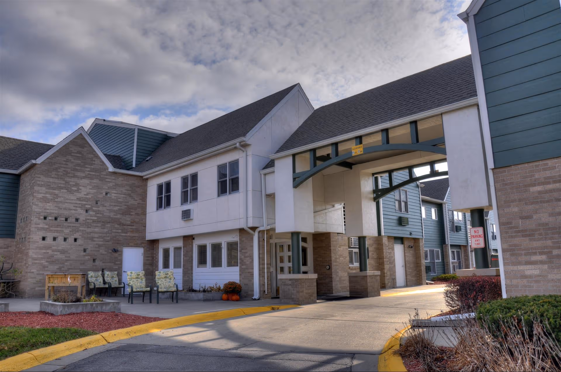 Exterior view of Hickory Villa Assisted Living facility showing a covered entrance with supporting pillars, multiple windows, and a seating area with chairs and a table. The building features a combination of brick and siding, with a cloudy sky overhead.