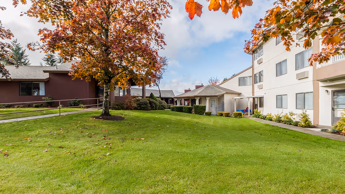 Outdoor view of Capital Place Retirement Community showing a well-maintained grassy lawn with a tree featuring autumn-colored leaves. The image includes parts of two buildings with multiple windows and a small covered entrance area under a partly cloudy sky.