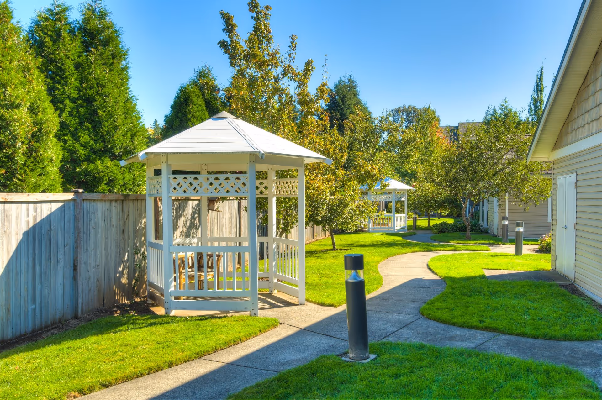 A sunny outdoor garden area with a paved walking path, green grass, trees, and two white wooden gazebos. There is a wooden fence on the left and a building on the right side of the image.