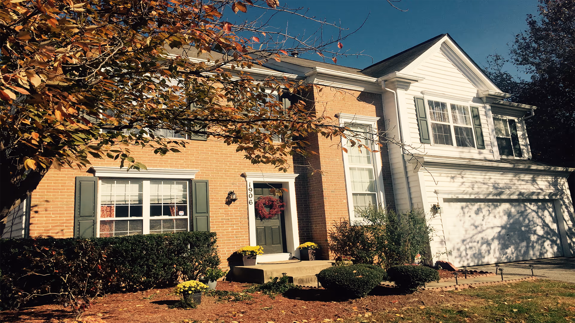 Exterior view of a two-story residential-style building with a brick and white siding facade, green shutters, a front door decorated with a wreath, and a garage. There are bushes and potted plants near the entrance, and a tree with autumn leaves partially shading the building.