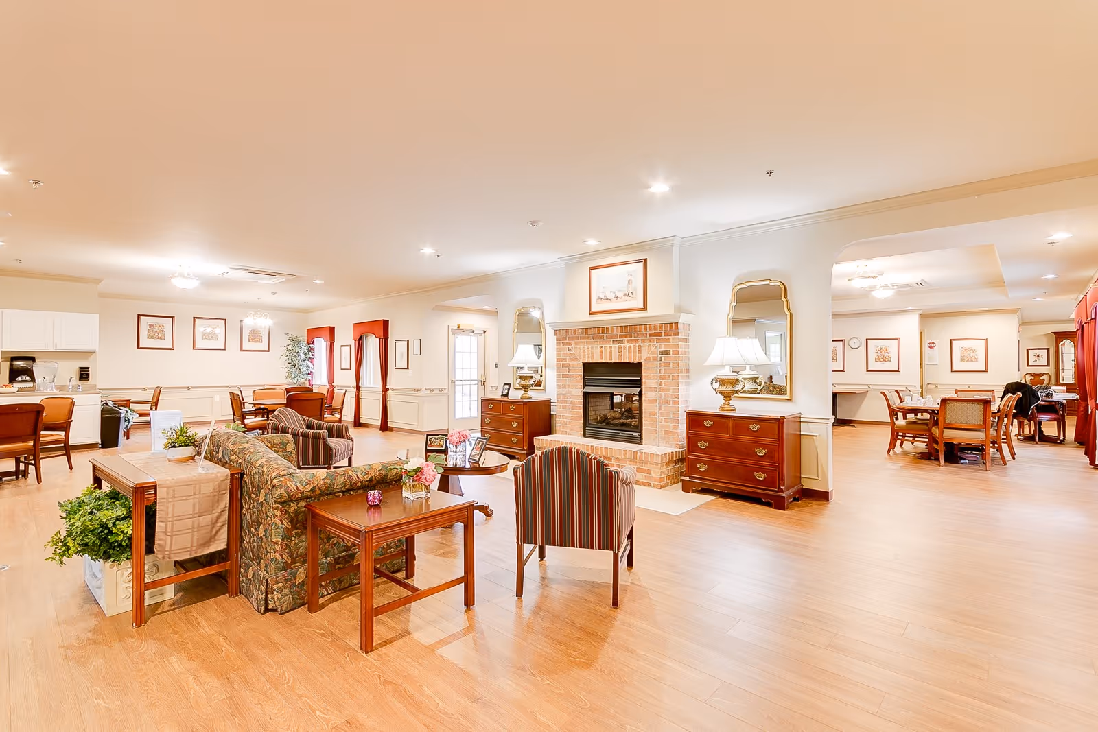 A spacious senior living common area featuring a brick fireplace flanked by two wooden dressers with lamps and mirrors above them. The room has multiple seating arrangements including floral and striped upholstered chairs and sofas, wooden tables with plants and flowers, and a dining area with tables and chairs in the background. The space is well-lit with ceiling lights and has wooden flooring and light-colored walls.