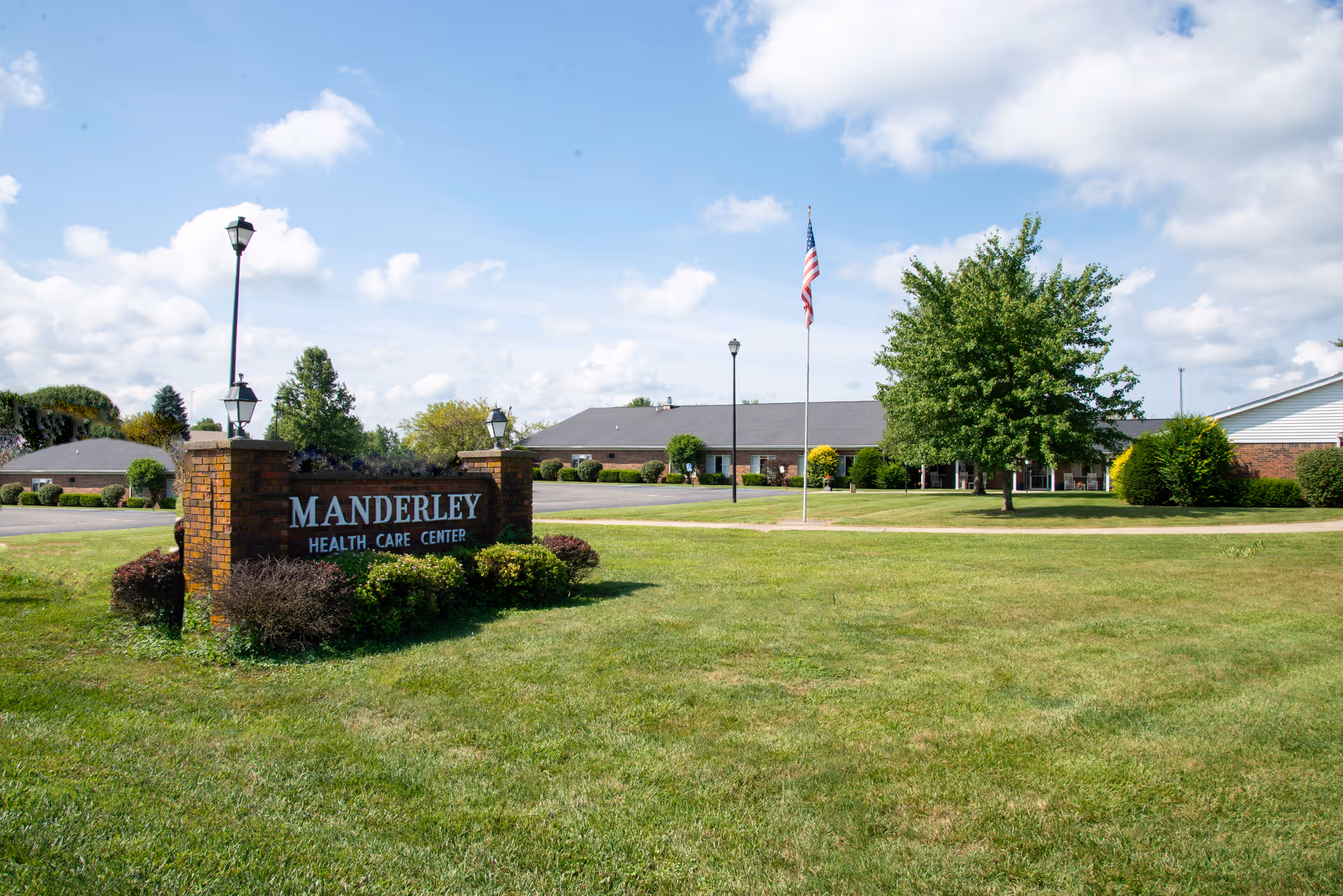 Brick sign reading 'MANDERLEY HEALTH CARE CENTER' on a grassy lawn with the facility building and an American flag in the background.