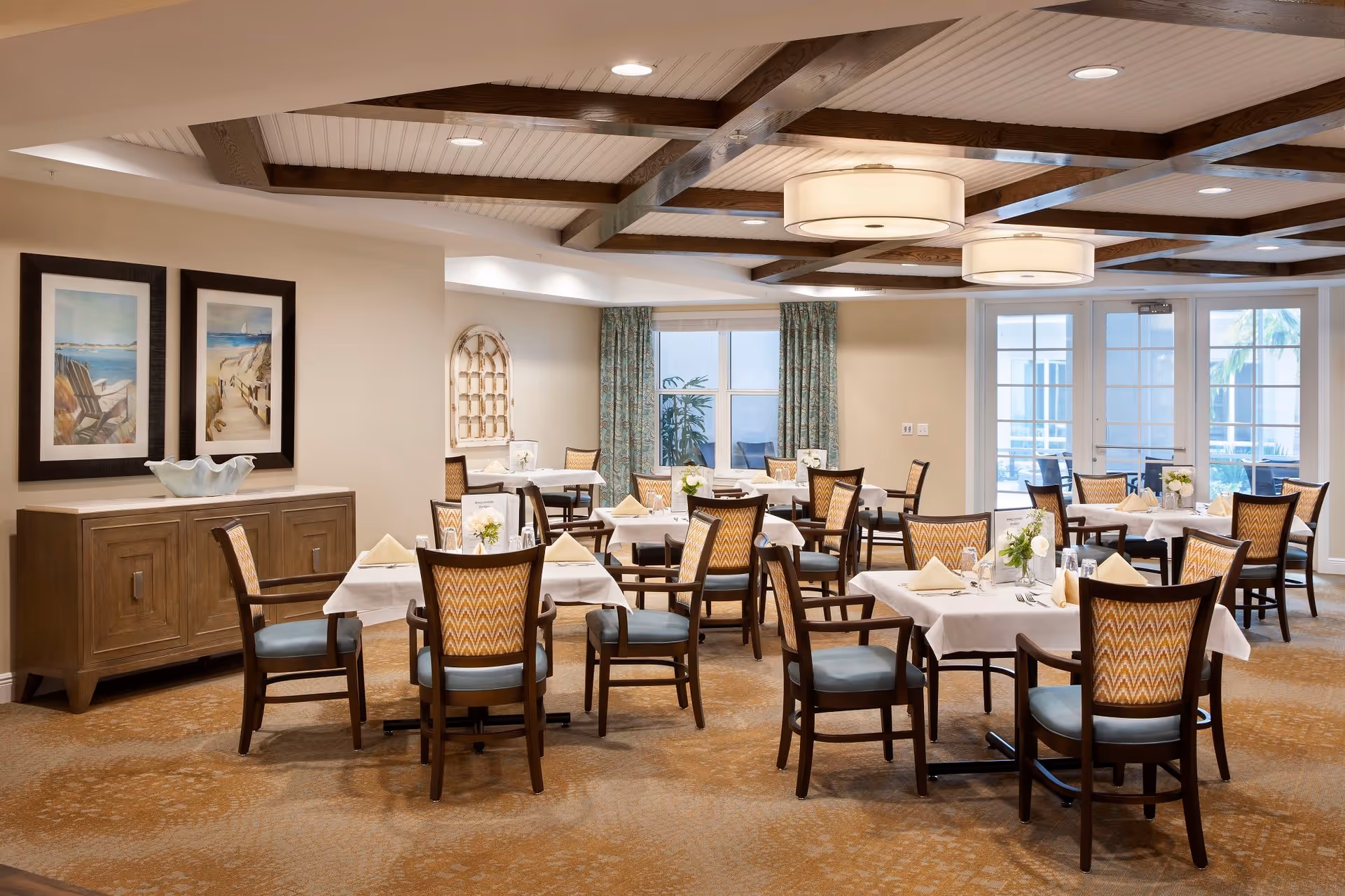 Bright, neatly arranged dining room with multiple set tables and wooden chairs under beamed ceilings.