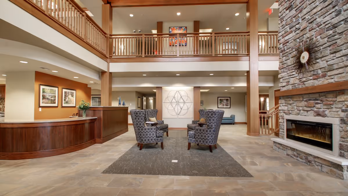 Spacious senior living facility lobby with a high ceiling and wooden balcony railing on the upper level. The room features a stone fireplace on the right, a wooden reception desk on the left, and two patterned armchairs facing each other on a rug in the center. The walls are decorated with framed artwork and a geometric wall sculpture.