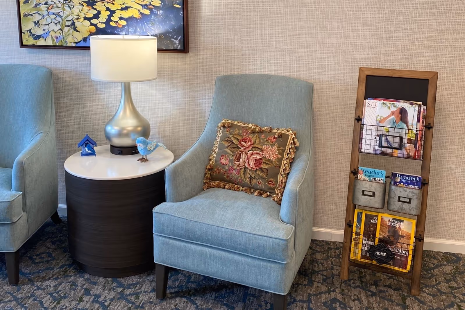 A cozy seating area with two light blue upholstered armchairs, one with a floral decorative pillow. Between the chairs is a round side table with a silver lamp and two small bird figurines. To the right is a wooden magazine rack holding various magazines including Reader's Digest and National Geographic. The wall behind has a textured beige wallpaper and a framed floral artwork.