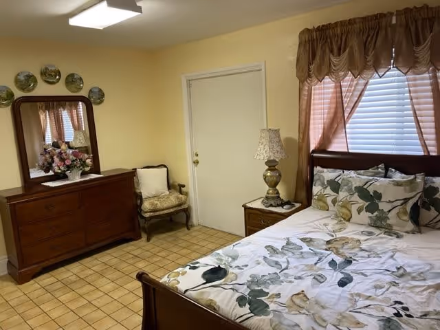 Bedroom with a floral-patterned bed, wooden headboard and dresser with mirror, nightstand with lamp, a chair, and tiled floor under soft yellow walls.
