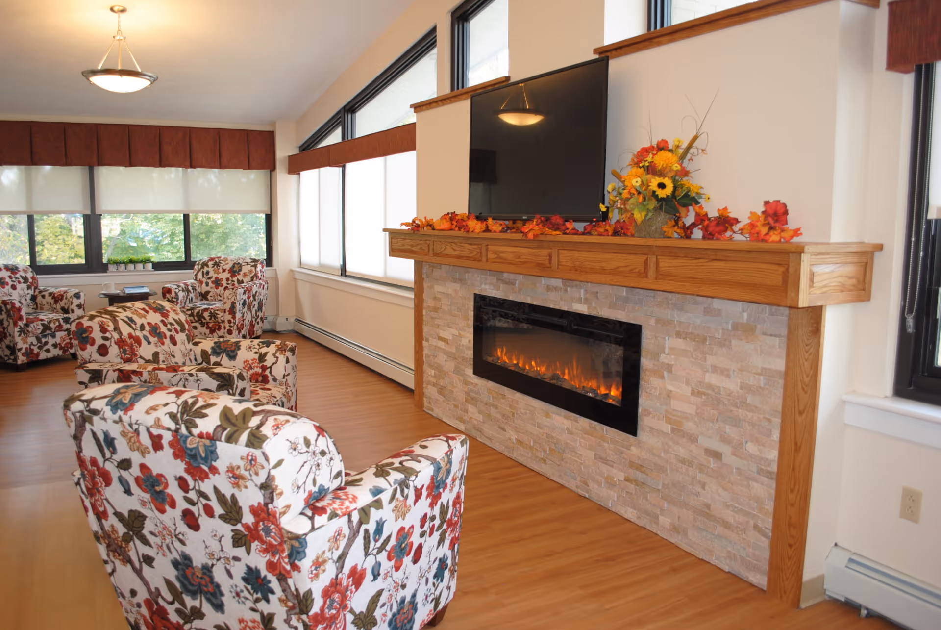 A cozy living room with floral patterned armchairs arranged around a modern electric fireplace set in a stone and wood mantel. Above the fireplace is a flat-screen TV mounted on the wall. The room has large windows with white blinds and red valances, allowing natural light to fill the space. A floral arrangement and autumn leaves decorate the mantel.