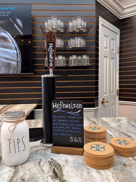 A countertop with a beer tap labeled Hefeweizen, a small chalkboard sign describing the beer, a jar labeled TIPS, and stacks of round cork coasters with a blue flower-like logo. Behind the counter are shelves holding multiple glass mugs, and a white door is visible in the background.