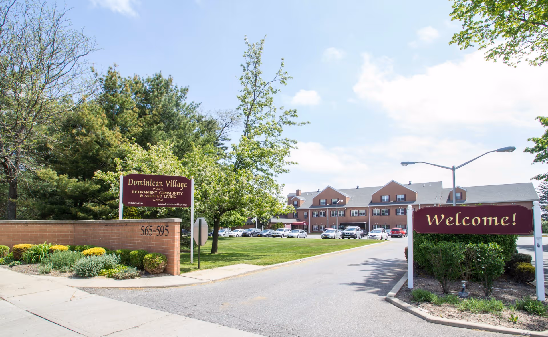 Entrance to Dominican Village retirement community and assisted living facility with a brick sign displaying the name and address, a 'Welcome!' sign, a driveway leading to a large building with multiple windows and parked cars, surrounded by green trees and landscaping under a partly cloudy sky.