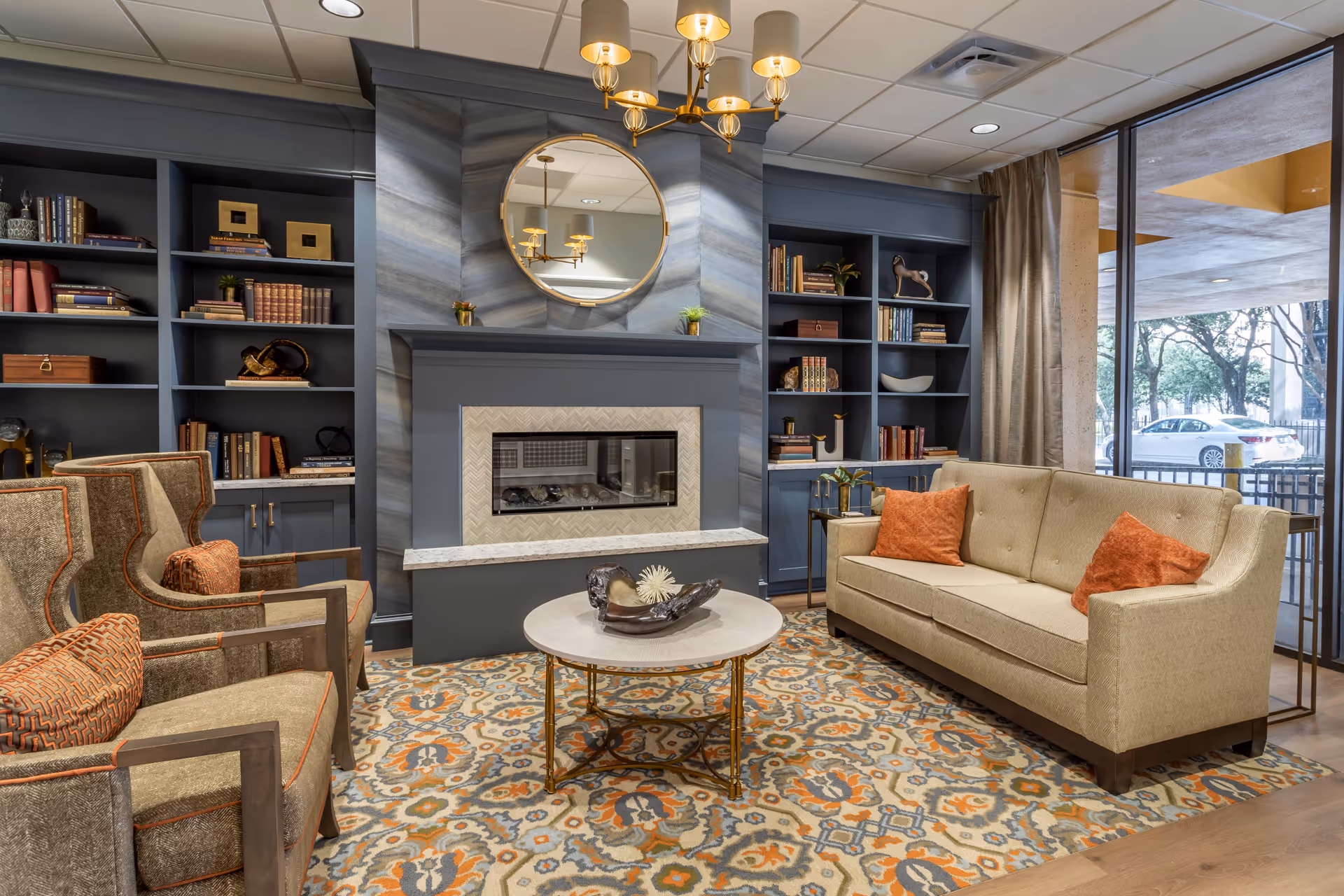 A cozy living room area with a beige sofa adorned with orange pillows, two armchairs with orange patterned cushions, a round coffee table with decorative items, a fireplace with a round mirror above it, built-in blue bookshelves filled with books and decor, and large windows showing an outdoor view with trees and a parked car.