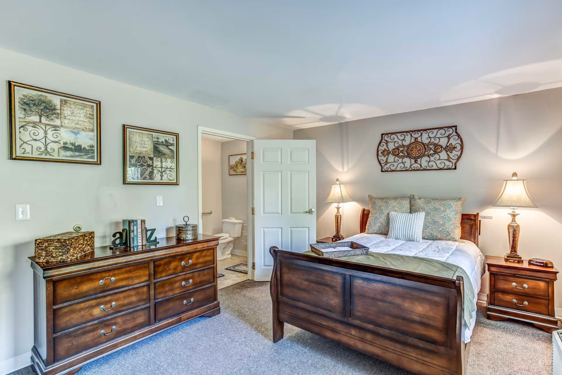 Well-lit bedroom with a wooden bed and matching dresser, nightstands with lamps, decorative wall art, and an open door to a bathroom.