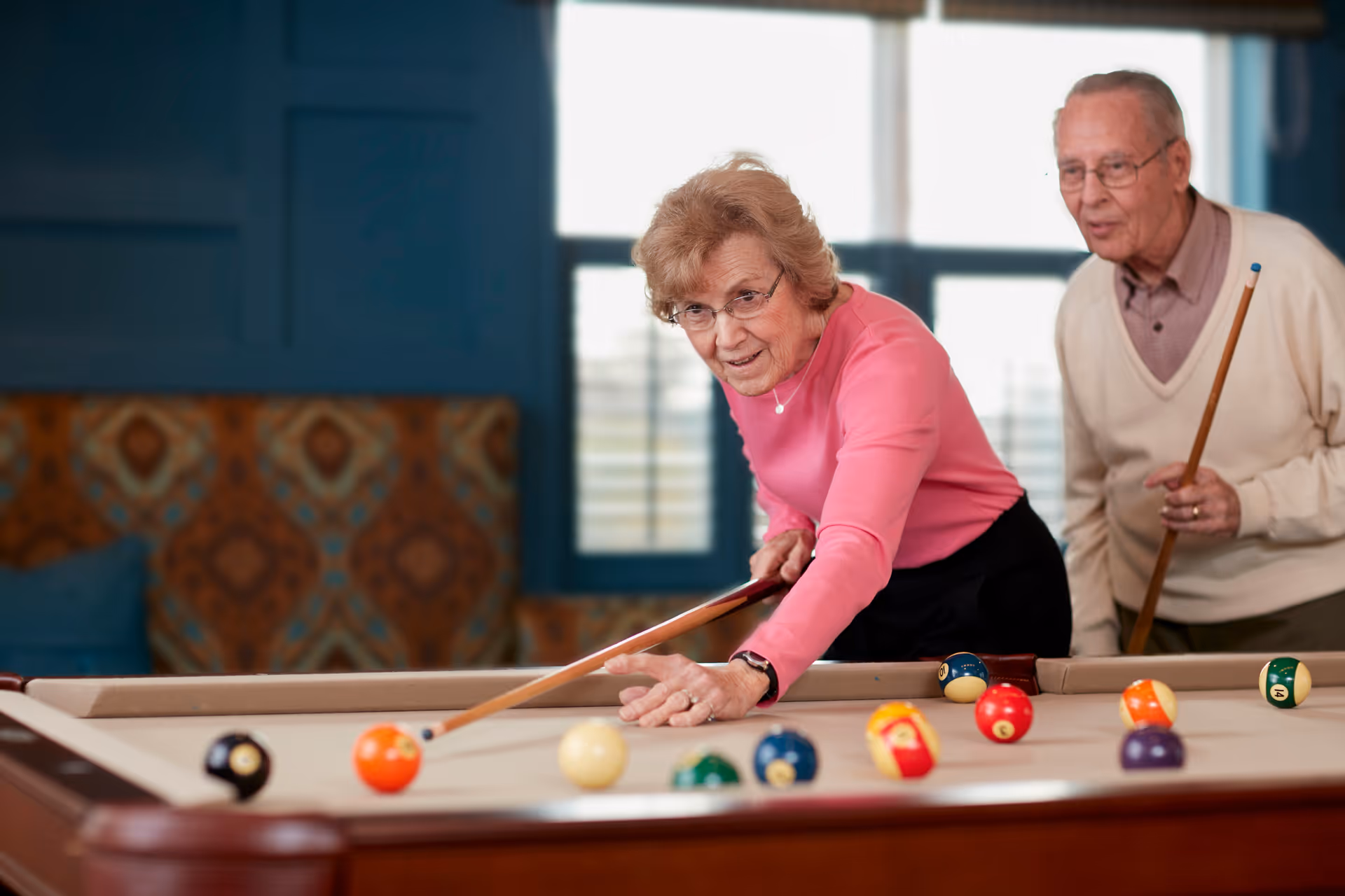 An elderly woman in a pink sweater is playing pool, aiming her cue stick at the cue ball on a billiards table, while an elderly man in a beige sweater stands behind her holding a cue stick, both inside a room with a window and patterned seating in the background.