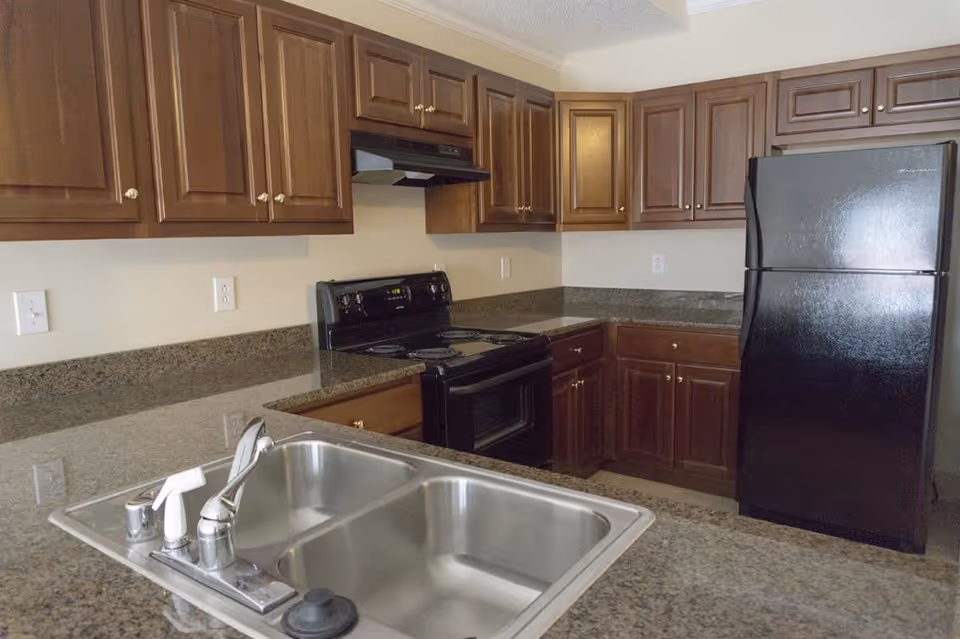 Kitchen area with dark wooden cabinets, granite countertops, a black electric stove with oven, a black refrigerator, and a double stainless steel sink with a faucet and soap dispenser.