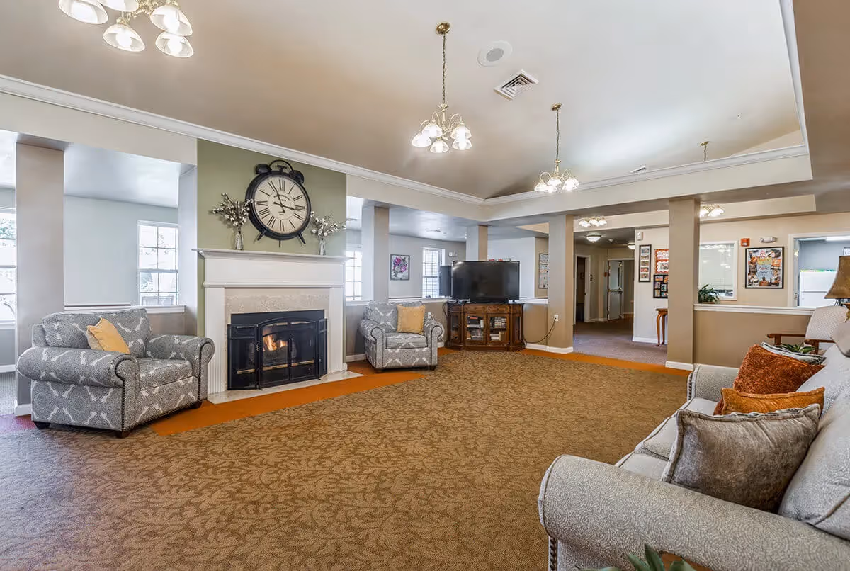 A spacious living room area with a large carpeted floor, two patterned armchairs with yellow pillows, a gray sofa with multiple cushions, a fireplace with a large clock above it, and a flat-screen TV on a wooden stand. The room has beige walls, multiple ceiling lights, and several windows letting in natural light.